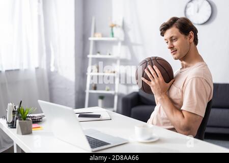 Concentration sélective de l'homme beau tenant le basket-ball près de l'ordinateur portable et de la papeterie sur la table à la maison, concept de gagner en ligne Banque D'Images