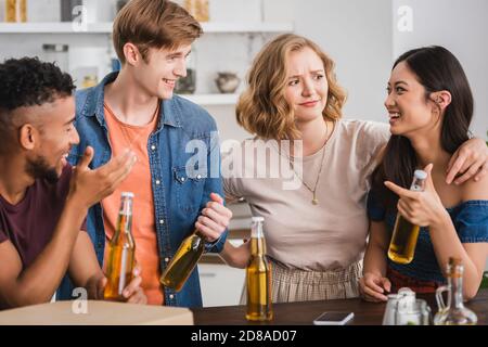 des amis multiculturels enthousiastes avec des bouteilles de bière qui parlent pendant la fête Banque D'Images