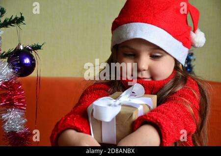 Une fille dans une petite casquette et un chandail décorent un arbre de Noël, ouvre des cadeaux, aime Noël, nouvel an. Bonne petite fille souriante avec cadeau de noël Banque D'Images