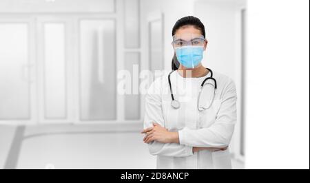 Vue de face d'une femme médecin de race mixte portant un écran de protection et un masque chirurgical, debout avec les bras croisés à l'hôpital de couloir Banque D'Images