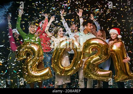Fête du nouvel an avec des amis. Groupe de jeunes heureux multiraciaux tenant des ballons de feuille d'or sous forme de nombres 2021 et dansant, confetti tombant dans l'air. Concept de fêtes et de célébrations Banque D'Images