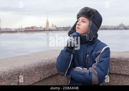 Adolescent habillé chaud veste et chapeau avec des rabats pensant, Pierre et Paul forteresse est sur un fond, pont de granit à Saint-Pétersbourg, Russie Banque D'Images