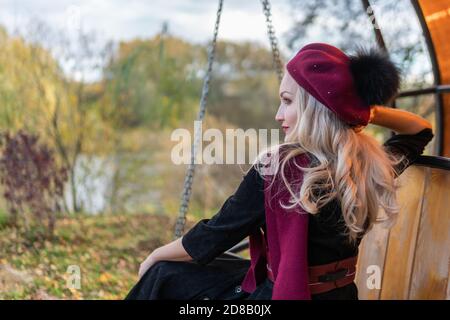 Une belle femme est assise sur une balançoire de jardin, forgée dans un manteau bordeaux et le biret, un adulte sourit à la caméra, à l'automne sur un fond de Banque D'Images