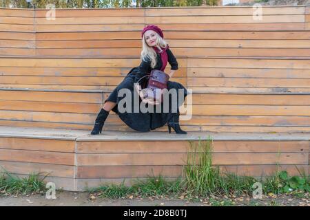 Happy s'assoit sur un banc d'un théâtre d'été, en bois dans un manteau bordeaux et le biret, regardant la caméra, à l'automne sur un fond de nuages bleus Banque D'Images