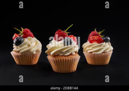 Trois petits gâteaux savoureux et sucrés dans la rangée, isolés sur fond noir. Vue rapprochée de l'avant d'un délicieux dessert frais avec garniture crémeuse blanche, tranches de fraises, de myrtilles et de baies de framboises. Banque D'Images