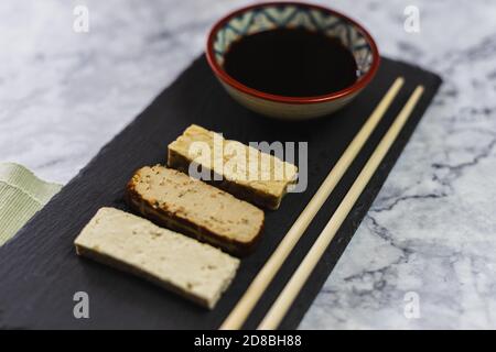 Table avec différents produits de soja. Tofu, tempeh et tofu fumé avec des graines. Bol avec sauce soja foncée et baguettes Banque D'Images
