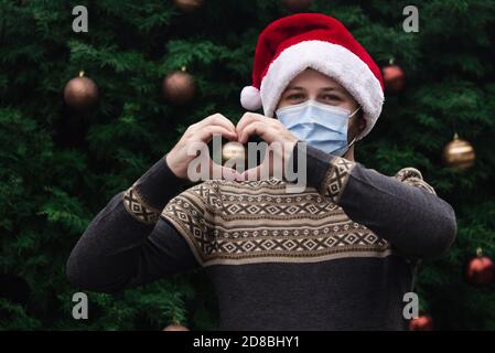 J'adore noël. Gros plan Portrait d'un homme portant un chapeau du père noël, un chandail de Noël et un masque médical avec émotion. Sur fond de Noël Banque D'Images