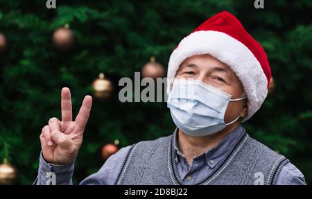 Article de Noël. Gros plan Portrait d'un homme âgé portant un chapeau du père noël et un masque médical avec émotion. Sur le fond d'un arbre de Noël. Banque D'Images