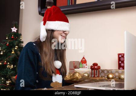 Chapeau de père Noël couvrant les yeux. Une jeune fille dans un chapeau de père noël et un chandail bleu se trouve près d'un ordinateur portable. La chambre est décorée avec goût. Noël pendant le TH Banque D'Images