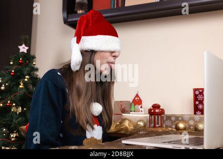 Chapeau de père Noël couvrant les yeux. Une jeune fille dans un chapeau de père noël et un chandail bleu se trouve près d'un ordinateur portable. La chambre est décorée avec goût. Noël pendant le TH Banque D'Images