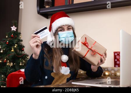 Achats en ligne de Noël. Une jeune fille dans un chapeau de père noël et un chandail bleu dans un masque médical se trouve près d'un ordinateur portable. La chambre est décorée avec goût. Banque D'Images