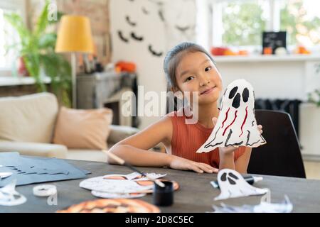 Portrait du contenu asiatique fille avec queue de cheval assis au bureau Avec des dessins d'Halloween et montrant l'artisanat fantôme à la caméra Banque D'Images