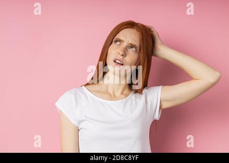 Pensive young caucasian ginger femelle avec des taches de rousseur peau cheveux longs portant des T-shirt blanc à la recherche jusqu'à l'expression perplexe, touchant ses lèvres de forefi Banque D'Images