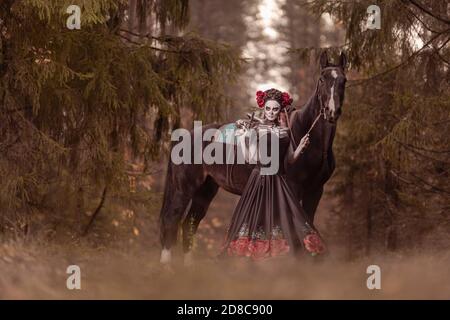 Jeune femme habillée comme le symbole mexicain de l'époque de la calavera morte dans une robe noire posant dans la forêt avec un cheval Banque D'Images