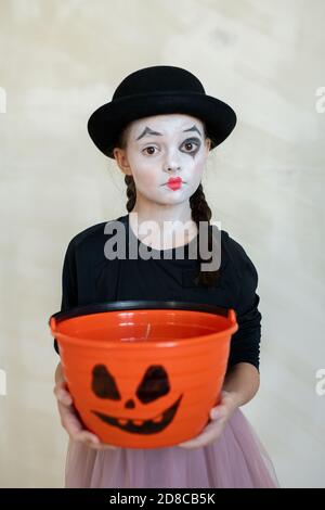 Portrait d'une fille mime choquée sérieuse dans un chapeau noir tenant un seau à traiter orange avec une image de visage effrayante sur un arrière-plan isolé, concept d'Halloween Banque D'Images