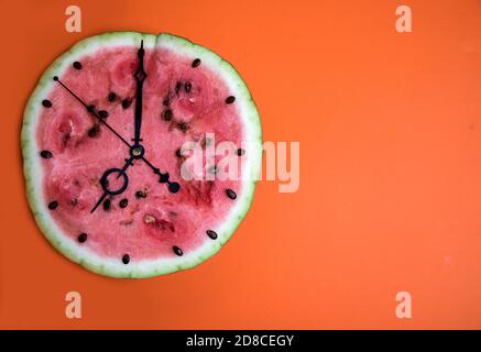 Tranche d'une pastèque mûre sous forme d'horloge, les mains montrent sept heures, fond orange. Orientation horizontale Banque D'Images