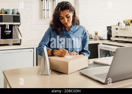 Jeune femme d'affaires debout dans son café préparant le colis avec le café pour l'expédition. L'entrepreneur écrit sur un autocollant sur une boîte en carton Banque D'Images