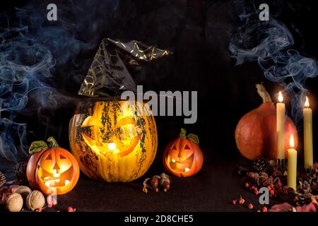 Citrouilles sculptées pour Halloween, la tête de Jack est sur la table. Citrouille dans un chapeau de sorcière. Fumer une bougie éclairée. Arrière-plan sombre. Banque D'Images