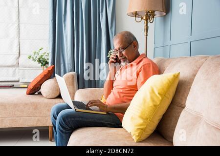 Portrait d'un homme indien asiatique moderne utilisant un ordinateur portable à tout en étant assis sur un canapé et en parlant sur un téléphone mobile ou un smartphone Banque D'Images