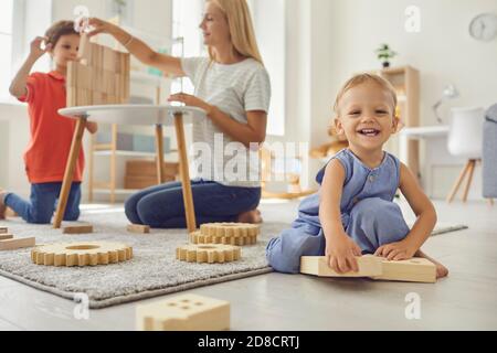 Mignon petit enfant regardant l'appareil photo et souriant pendant l'aîné frère et maman jouent en arrière-plan Banque D'Images