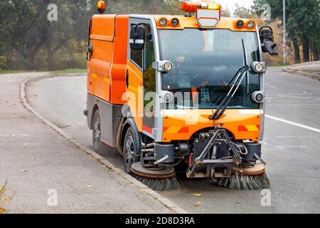 Un petit balai de rue avec deux brosses avant réglables indépendamment et à réglage hydraulique nettoie la route dans un quartier résidentiel de la ville. Banque D'Images