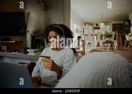 Jeune femme africaine avec carte de crédit à la main assise avec ordinateur portable et transfert d'argent et écoute de musique Banque D'Images