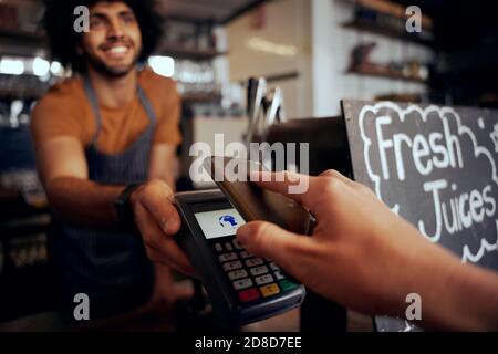 Gros plan de la main jeune femme effectuant le paiement sans contact dans le café utilisation d'un smartphone avec un serveur tenant la machine Banque D'Images