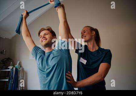 Vue à angle bas d'un jeune homme souriant faisant des exercices avec beau jeune thérapeute utilisant une bande élastique Banque D'Images