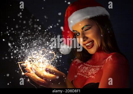 Une belle fille brune avec maquillage d'art, portant un chapeau de Père Noël, se tient souriant sur un fond bleu dans le studio Banque D'Images