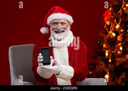 Portrait du Père Noël barbu en lunettes souriant à l'appareil photo tenir le téléphone portable dans ses mains Banque D'Images