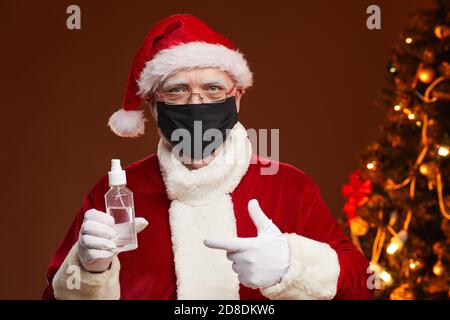 Portrait du Père Noël en costume rouge portant un masque de protection pointant vers l'antiseptique dans sa main debout au studio Banque D'Images