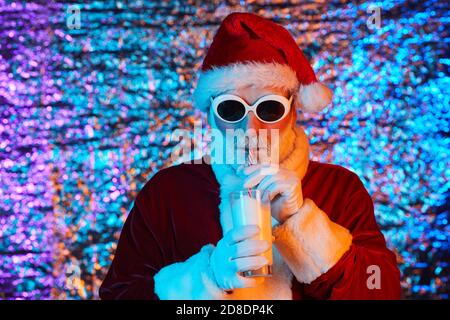 Le Père Noël dans des lunettes de soleil tenant le verre et de boire du lait de boire de la paille sur un fond brillant Banque D'Images