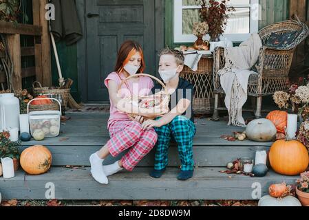 enfants un garçon et une fille en pyjama et de protection masque facial avec un panier de bonbons sur le porche de l'arrière-cour décoré avec des citrouilles en automne Banque D'Images