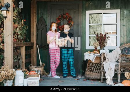 enfants un garçon et une fille en pyjama et de protection masque facial avec un panier de bonbons sur le porche de l'arrière-cour décoré avec des citrouilles en automne Banque D'Images