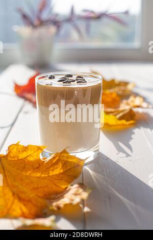 Boisson traditionnelle au lait caféiné coréen, café Dalgona dans un verre avec des grains de café sur fond de tableau blanc et une fenêtre. Feuilles d'érable sèches. Au Banque D'Images