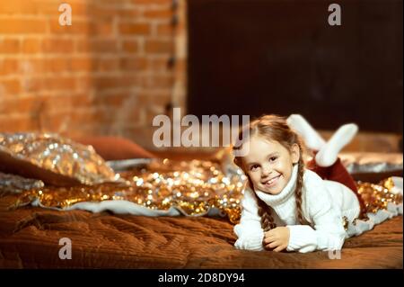 petite fille dans une robe de noël. la fille se trouve sur le lit et sourit. Photo confortable de la maison. Le confort de Noël à la maison Banque D'Images