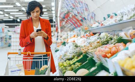 Au supermarché: Belle jeune femme marche à travers la section des produits frais, choisit des légumes et pousser le panier. Le client utilise un smartphone Banque D'Images