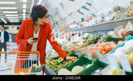 Au supermarché: Belle jeune femme marche à travers la section des produits frais, choisit des légumes et les place dans son panier. Le client utilise Banque D'Images