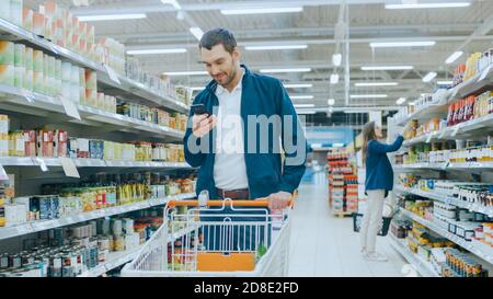 Au supermarché: Handsome Man utilise smartphone, Smiles tout en se tenant à la section des marchandises en conserve. Contient un panier contenant des produits alimentaires sains Banque D'Images