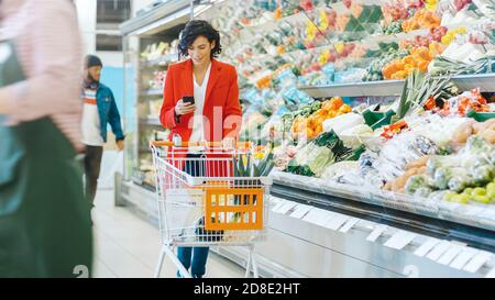 Au supermarché: Belle jeune femme avec panier utilise un smartphone et marche à travers la section des produits frais du magasin. Banque D'Images