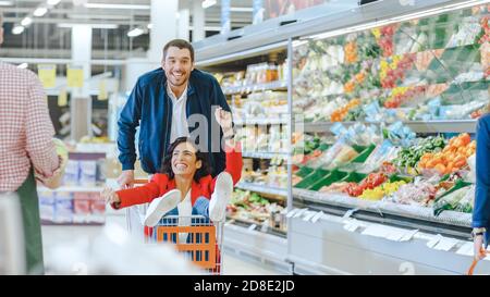 Au supermarché : l'homme pousse le chariot avec une femme assise dans celui-ci. Happy couple a de l'amusement pendant que course sur un tramway à travers les produits frais Banque D'Images