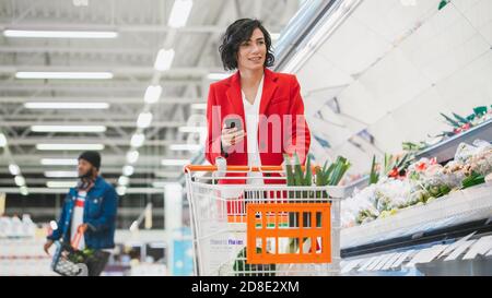 Au supermarché: Belle jeune femme avec panier utilise un smartphone et marche à travers la section des produits frais du magasin. Banque D'Images