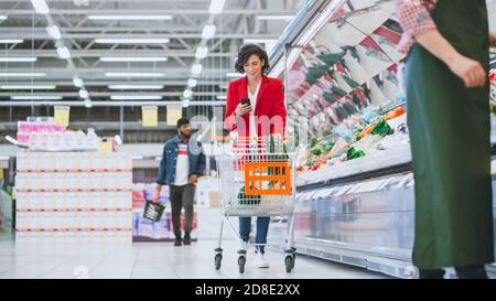 Au supermarché: Belle jeune femme avec panier utilise un smartphone et marche à travers la section des produits frais du magasin. Banque D'Images