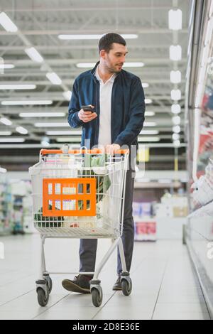 Au supermarché : la prise de vue verticale complète d'un jeune homme charmant utilise un smartphone tout en poussant le panier et en choisissant des légumes frais Banque D'Images