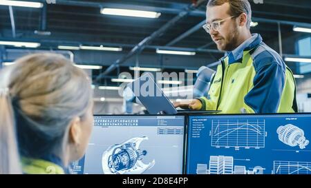 À l'usine : les ingénieurs mécaniques hommes et femmes utilisent un ordinateur portable et un ordinateur personnel tout en discutant du projet de fabrication de bras robotiques. L'écran s'affiche Banque D'Images