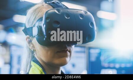 À l'usine : une femme ingénieure portant un casque de réalité virtuelle travaille sur un projet, déplaçant sa tête. Dans les écrans arrière-plan et industriel Banque D'Images