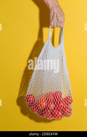 Main mâle tenant un sac en maille blanche avec des légumes sur fond jaune avec un espace de copie. Concept de shopping zéro déchet. Sac à corde avec tomatoe Banque D'Images