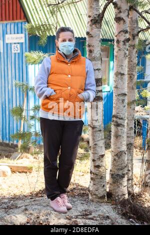 Femme de campagne portant un masque médical et un gilet orange bouleaux pendu près de sa maison d'été, auto-isolation dans un village Banque D'Images