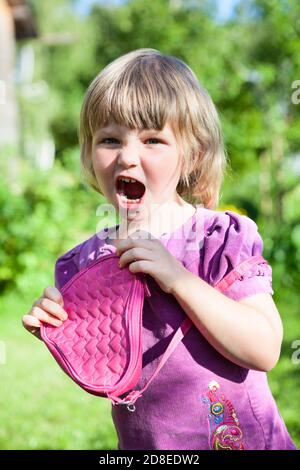 Petite fille caucasienne criant avec un petit sac rose dans les mains, portant la robe dans le jardin d'été, enfant en colère Banque D'Images