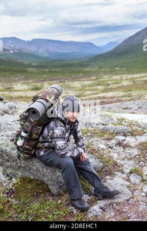 Adulte alpiniste caucasien reposant sur la pierre avec sac à dos lourd sur son dos, homme randonnée dans la vallée de montagne Banque D'Images